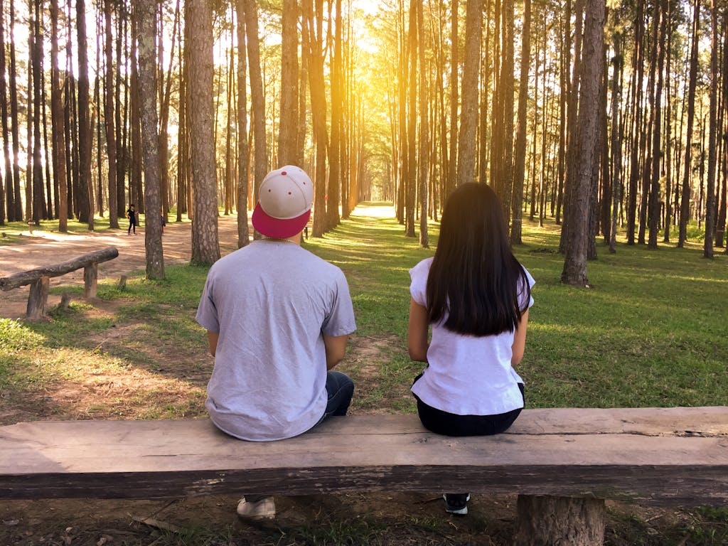 A young couple sits on a bench surrounded by tall trees in a forest at sunset.